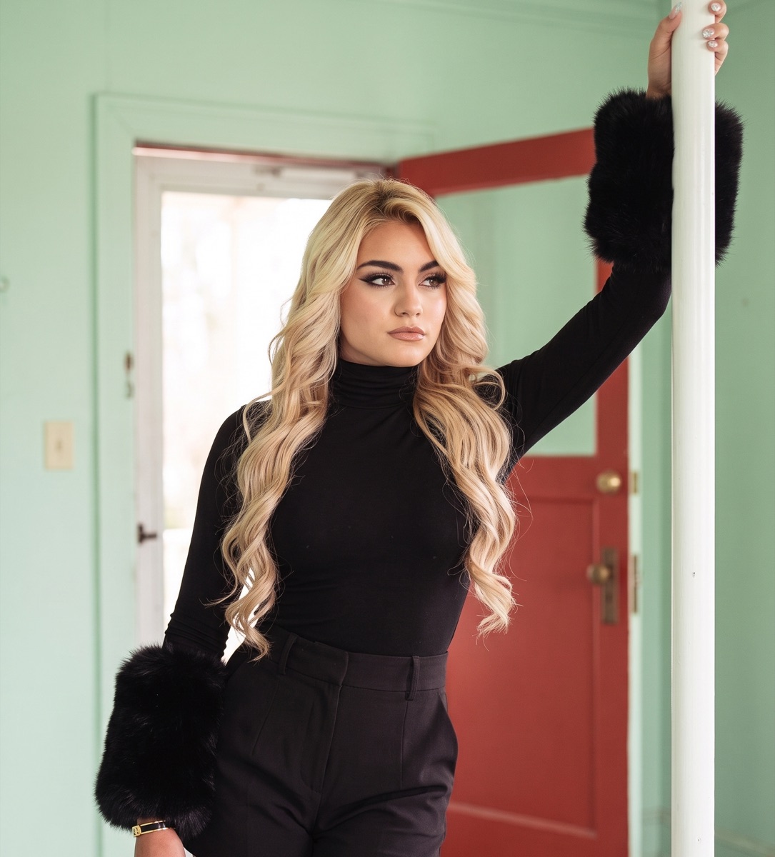 SUPPA in a black turtleneck with fur cuffs in a mint-green room